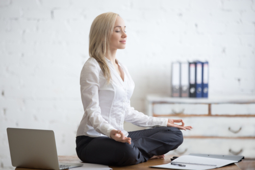 Business and healthy lifestyle concept. Portrait of young office woman sitting cross-legged in half lotus yoga pose at workplace. Smiling business lady meditating after finishing her work
